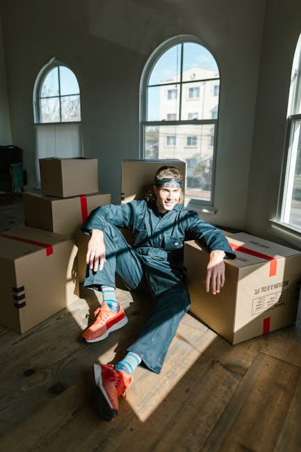 A young woman with curly black hair and a cheerful expression is sitting on a wooden floor surrounded by numerous stacked cardboard moving boxes of various sizes, some sealed with packing tape and oth