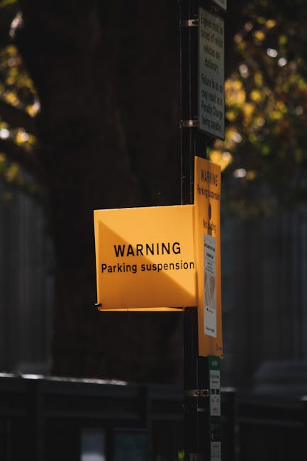 A tall, rectangular 'No Parking' sign with a white background and red border, mounted on a grey metal pole, is positioned on a paved sidewalk. The sign features bold red lettering that reads 'NO PARKI