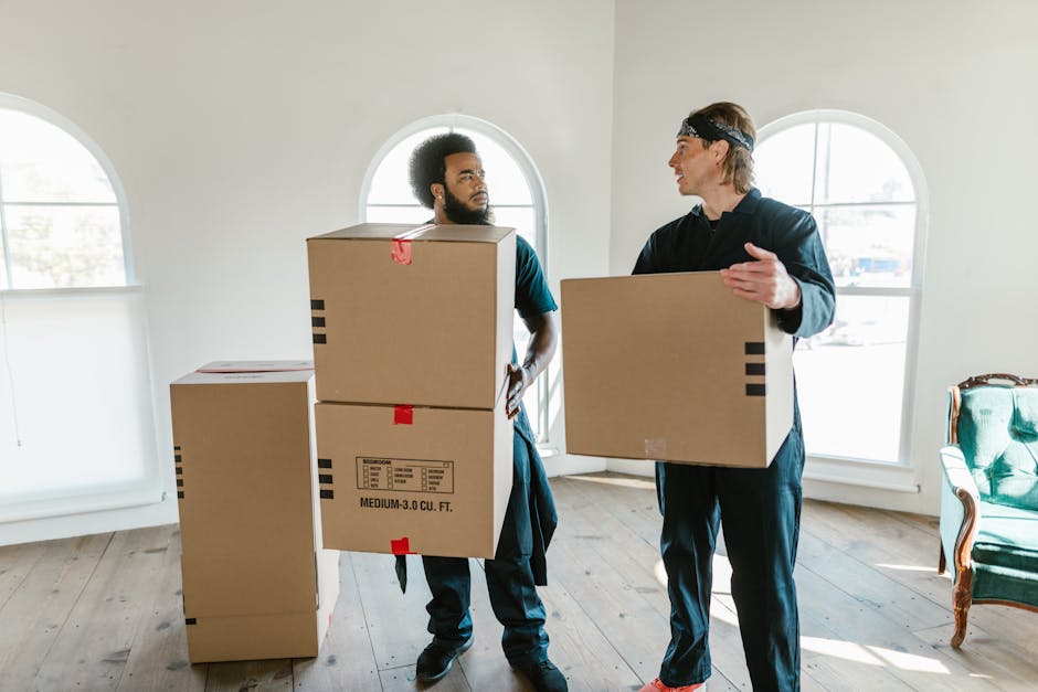 A young man with light skin, wearing a blue work uniform, a headscarf, and red-orange sneakers, is sitting on a wooden floor inside a room with large, arched windows that let in natural light. He is s