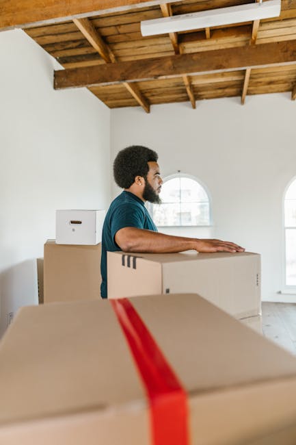 A person, whose torso and arms are visible, is engaged in packing or preparing for a house move inside a well-lit room with large windows. They are handling a cardboard box, which they are placing on